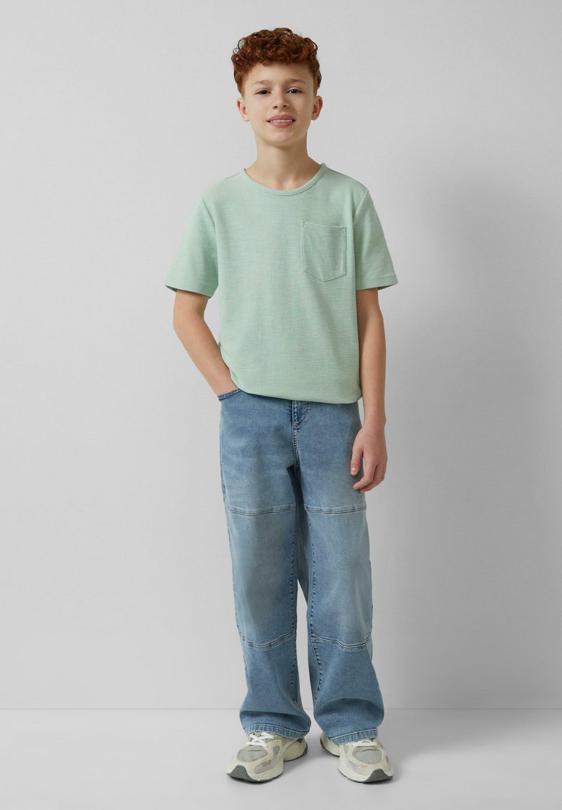 Lichtgroene t-shirt met korte mouwen en een zak, gecombineerd met lichtblauwe denim broeken met een patchwork patroon en neutrale sneakers.