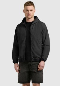 Muž v čiernej mikine s kapucňou na zips a čiernych rifľových kraťasoch stojaci pred jednofarebným pozadím.