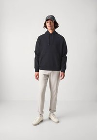 Fekete kapucnis pulóver elülső zsebbel és húzózsinórral; világosszürke keskenyedő nadrág; szürke sapka; fehér, mintás talpú sportcipő. Semleges háttér.