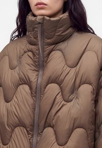 Femme portant une doudoune marron matelassée avec un col montant et une fermeture éclair visible à l'avant, montrant le haut du torse et le cou.
