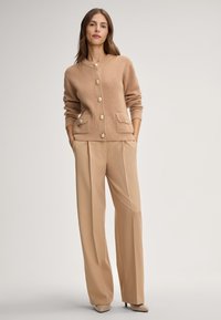 Donna che indossa un cardigan a coste beige con bottoni dorati e pantaloni a gamba larga coordinati, in piedi con le mani in tasca su uno sfondo neutro.