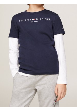 Barn iført en mørkeblå Tommy Hilfiger t-shirt over hvide langærmede trøjer og lysegrå joggingbukser med logo på låret.