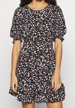 Kobieta nosząca czarną sukienkę w kwiaty w kolorach różowym, białym i czerwonym, z bufkami na krótkich rękawach i falbaną u dołu.