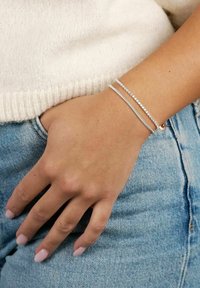 Silverarmband med två strängar: en enkel och en med små klara stenar, bärs på handleden. Handen vilar mot denimtyg.