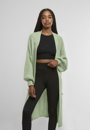 Langer, offener Strickcardigan in sanftem Hellgrün, mit einem entspannten Schnitt, überschnittenen Schultern und gerippter Textur. Kombiniert mit einem schwarzen Cropped-Top und Leggings.