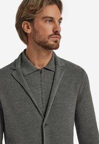 Grauer Strickcardigan aus strukturiertem Rippstoff, mit schmalem Kragen und einem Reißverschluss. Verfügt über eine Knopfleiste mit zwei Knöpfen und hat ein weiches Finish.