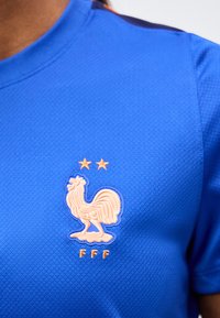 Blau texturiertes Sporttrikot mit erhabenem orangefarbenem Hahn-Emblem, zwei Sternen darüber und den Buchstaben "FFF" darunter auf der Brust.