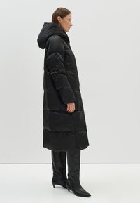 Zwarte pufferjas met capuchon, met een lang, doorgestikt ontwerp en een gladde textuur. Gecombineerd met zwarte leren laarzen tot de knie.