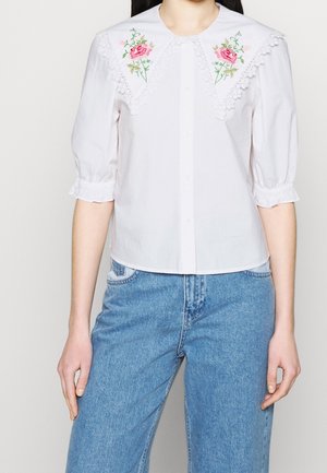Blouse blanche avec grand col festonné brodé de roses roses, manches bouffantes jusqu'au coude, et jean bleu taille haute.