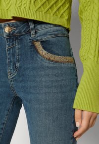 Jeans de mezclilla azul con un patrón de costura detallado en el bolsillo. Combinados con un suéter de punto texturizado en un verde brillante.