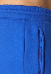 Calções atléticos azuis com uma textura suave, cintura elástica e um bolso com fecho lateral para armazenamento. Apresentam um acabamento liso e uniforme.