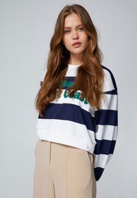 Sudadera a rayas en blanco y navy con texto bordado en verde, que presenta un corte holgado y mangas amplias. Unos pantalones beige de cintura alta completan el conjunto.