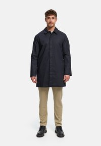 Navy dugi kaput sa šiljastim ovratnikom i zatvaračem na kopčanje, dizajniran s ravnim krojem. Nošen preko bež hlača i crnih cipela.