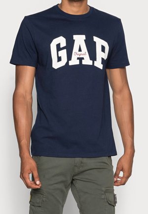 T-shirt in cotone blu navy con la parola "GAP" stampata in grandi lettere bianche, con "Original" in rosso sottostante, scollatura rotonda, maniche corte.