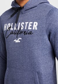 Marinblå hoodie i mjukt tyg, med dragsko på huvan, stor framficka och vit broderad text "HOLLISTER California".