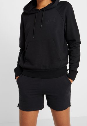 Persoon die een zwarte capuchontrui met voorzak en een zwarte korte broek draagt, handen in de zakken, staand tegen een effen achtergrond.