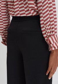 Zwarte op maat gemaakte broeken met een gladde textuur en twee achterzakken, gecombineerd met een rood-wit gestreepte blouse met knoopsluiting bij de mouwen.