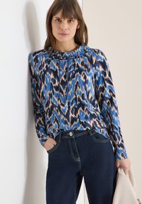 Femme portant un haut à manches longues bleu, noir et beige à motifs, avec un jean en denim foncé, tenant une veste beige clair, debout contre un mur blanc.