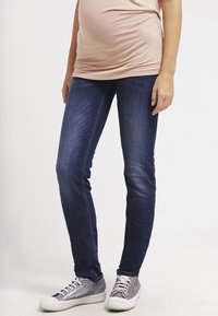 Calças skinny de denim em azul escuro, com uma textura suave e desbotado leve, combinadas com ténis cinzentos e uma blusa ajustada rosa.