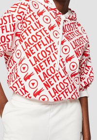 Sweat à capuche blanc avec texte rouge répété « LACOSTE NETFLIX », logos crocodile rouges et icônes de bouton lecture rouges, porté avec un pantalon blanc.