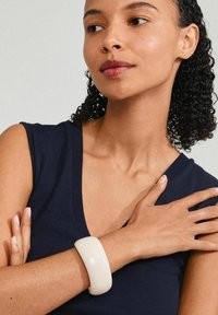 Biely lesklý náramok na zápästí, ktorý vystavuje hladký, zaoblený dizajn. Modelka má na sebe tmavomodrý top bez rukávov.