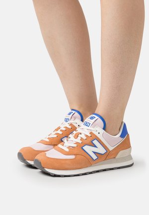 Jambes portant des baskets New Balance 574 orange, blanches et bleues avec des lacets blancs sur un fond blanc.