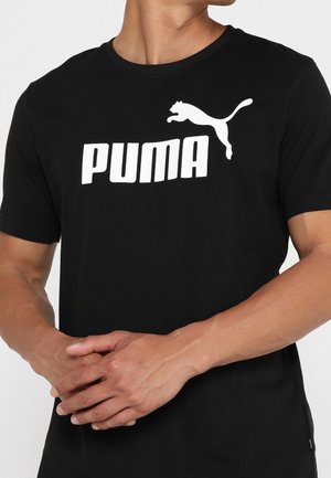 Personne portant un t-shirt noir Puma avec un logo Puma blanc et un texte sur le devant, mains jointes devant.