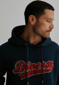Granatowy hoodie z dużym, wyhaftowanym w kolorze czerwonym i kremowym logo "Diversity". Sznurki są czerwone z metalowymi końcówkami, widoczna tekstura bawełny.