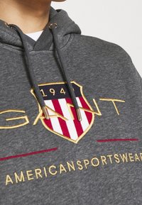 Felpa grigia con cappuccio, caratterizzata da un grande emblema con strisce rosse e bianche, testo ricamato "GANT" e "AMERICAN SPORTSWEAR" in oro. Fettuccia regolabile nel cappuccio.