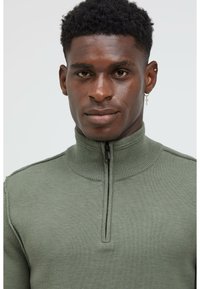 Khaki-grüner Zip-Pullover aus geripptem Strickmaterial; verfügt über Nähte und einen hohen Kragen für zusätzliche Wärme.