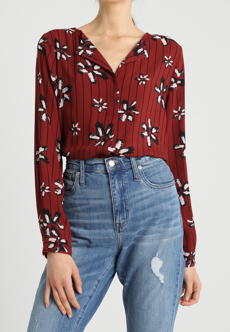 Blusa rossa floreale con maniche lunghe, scollatura a V e righe verticali, abbinata a jeans in denim blu a vita alta con dettagli strappati.