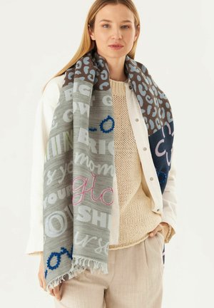 Grau und braun gemusterter Schal mit Text in verschiedenen Farben, mit fransigen Kanten. Getragen über einem hellen Pullover und einer Jacke.