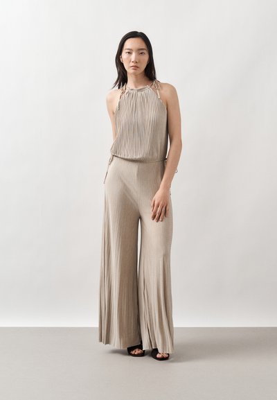 Femme portant un haut halter plissé beige et un pantalon à jambes larges assorti, debout devant un fond clair uni.