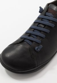 Zapato de cuero negro con cordones azul marino. Punta redondeada y detalles cosidos a lo largo del lateral. Textura suave con acentos mínimos.