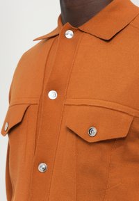 Camisa naranja de canalé con collar puntiagudo, dos bolsillos en el pecho y botones a presión plateados para el cierre.