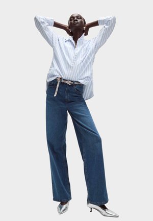 Blau-weiß gestreiftes Hemd mit Knöpfen, lockere Passform. Dunkelblaue Jeans mit weitem Bein. Silberne, spitze High Heels. Glitzerndes Detail am Gürtel. Schlichter Hintergrund.