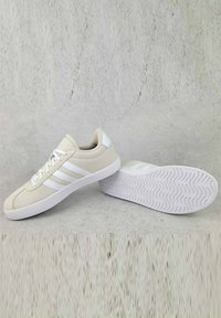 Scarpe da ginnastica bianche con un upper liscio, caratterizzate da tre strisce bianche e una suola bianca texturizzata con un motivo ondulato.