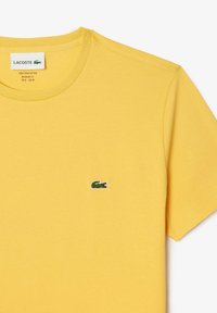 Geel katoenen T-shirt met een ronde hals en korte mouwen. Heeft een klein groen Lacoste krokodil logo op de linkerborst.