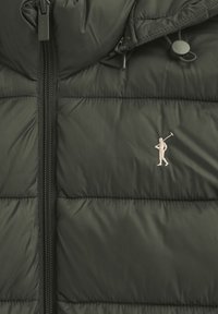 Mörkgrön quiltad jacka med dragkedja framtill och en liten broderad figur som håller i en golfklubba på bröstet.