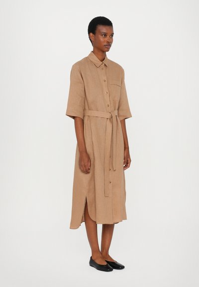 Vestido longo, cor de camelo, com botões, colarinho, mangas curtas, uma fenda lateral e uma cintura cingida; usado com sapatos pretos planos.