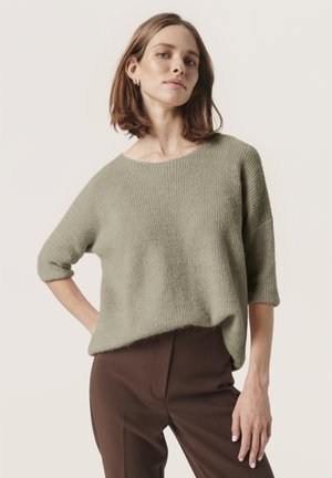Lichtgroene, geribbelde gebreide trui met een wijde halslijn en driekwartmouwen, gecombineerd met een bruine broek. Zachte textuur en een relaxed fit.