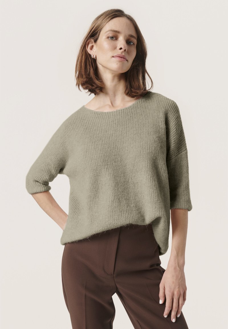 Lichtgroene, geribbelde gebreide trui met een wijde halslijn en driekwartmouwen, gecombineerd met een bruine broek. Zachte textuur en een relaxed fit.