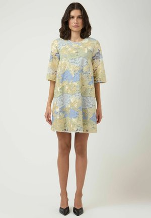 Vrouw staand, draagt een losse, knielange kanten jurk met gele en blauwe bloemenpatronen, gecombineerd met zwarte puntige hakken.