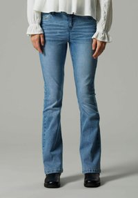 Niebieskie dżinsy z rozszerzanymi nogawkami, wykonane z denimu o jasnym kolorze, standardowy pięciokieszeniowy krój, lekkie przetarcia i noszone z czarnymi butami.