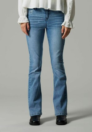 Zilas platās džinses, izgatavotas no denima ar gaiši zilu mazgāšanu, standarta piecu kabatu dizains, neliela nolietojuma pazīmes un valkātas ar melnām zābakiem.