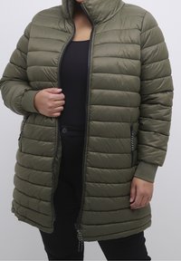 Oliivinvihreä toppatakki, jossa vaakasuuntaiset tikatut kuviot, korkea kaulus, vetoketju edessä ja sivutaskut, sekä resorireunukset ja teksturoitu kangas.