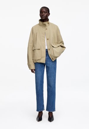 Femme noire portant une veste beige surdimensionnée boutonnée, un jean bleu droit, des chaussures noires pointues et un haut blanc sur un fond blanc.