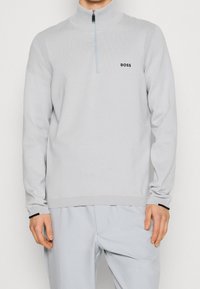 Pull demi-zippé gris clair en tissu doux, présentant un logo noir sur la poitrine et des accents côtelés le long des épaules et des poignets.