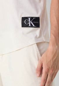 Fehér pamut póló, fekete hímzett címkével, rajta "CK" és "Calvin Klein Jeans" felirattal. Puha tapintású és laza szabású.