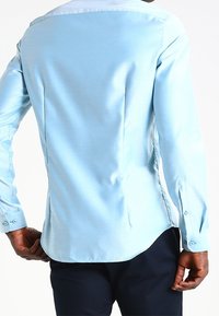 Camisa de manga larga azul claro con tela suave, corte ajustado, dobladillo curvado y puños abotonados. La parte trasera presenta costuras estructuradas.